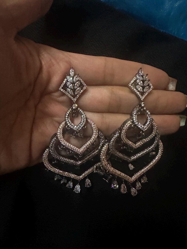May include: A pair of ornate, silver-toned chandelier earrings. Each earring features a diamond-shaped top with a leaf design, followed by layered, heart-shaped tiers, and teardrop-shaped dangles. The earrings are set with clear, sparkling stones.