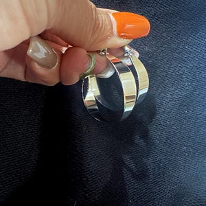 Op de afbeelding: Een paar zilveren oorringen. De oorbellen zijn dik en glanzend en reflecteren licht. De ringen worden vastgehouden door een hand met oranje nagellak. De achtergrond is een donkere, getextureerde stof.