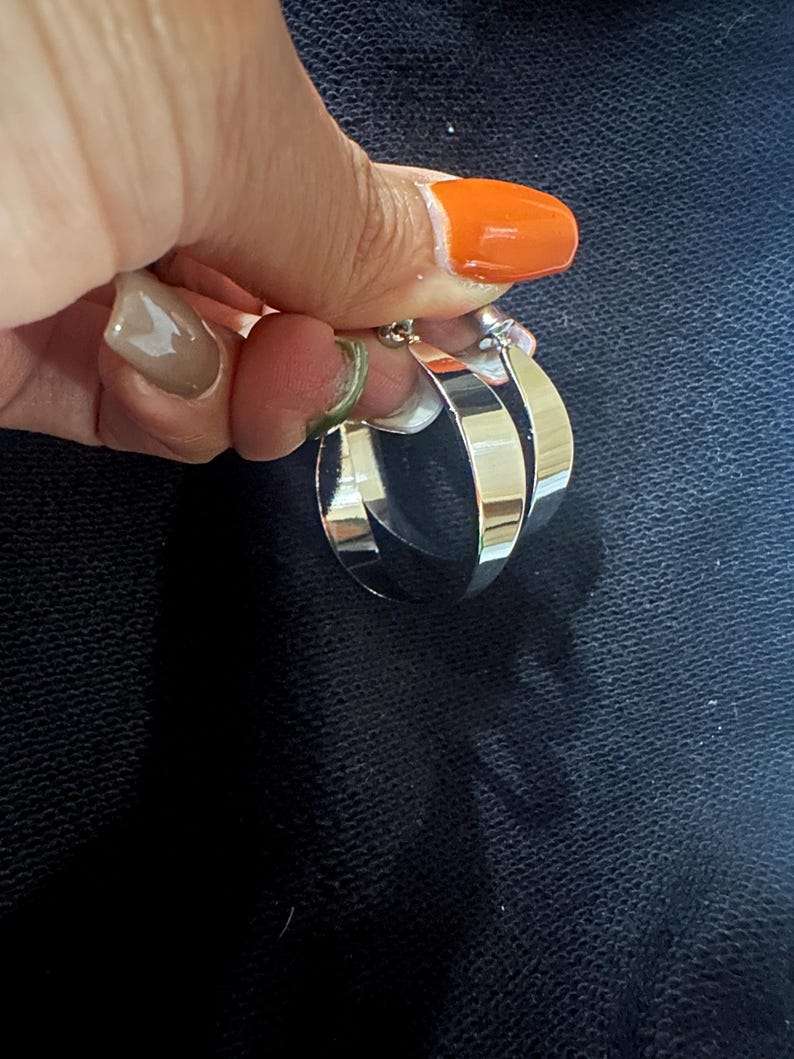 Op de afbeelding: Een paar zilveren oorringen. De oorbellen zijn dik en glanzend en reflecteren licht. De ringen worden vastgehouden door een hand met oranje nagellak. De achtergrond is een donkere, getextureerde stof.