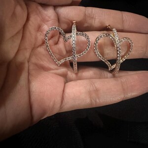 Pu&ograve; includere: Un paio di orecchini a cerchio color oro, ciascuno a forma di cuore e incrostato di piccoli cristalli scintillanti. Gli orecchini sono tenuti in una mano, mostrando il loro design delicato e i dettagli intricati. Il design a forma di cuore aggiunge un tocco romantico.