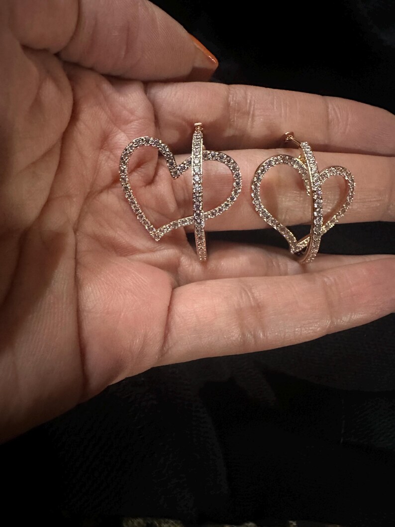 Pu&ograve; includere: Un paio di orecchini a cerchio color oro, ciascuno a forma di cuore e incrostato di piccoli cristalli scintillanti. Gli orecchini sono tenuti in una mano, mostrando il loro design delicato e i dettagli intricati. Il design a forma di cuore aggiunge un tocco romantico.