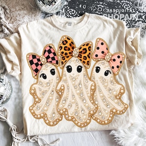 May include: A cream-colored t-shirt featuring a graphic of three ghosts adorned with white pearls and clear crystals. Each ghost wears a large bow: one with a pink and black checkerboard pattern, one with a leopard print, and one with pink polka dots.