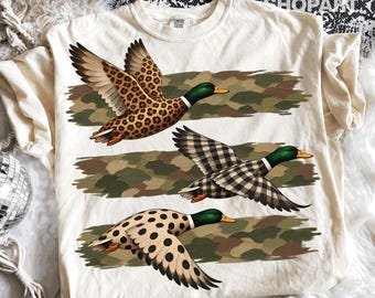 May include: A cream-colored t-shirt featuring three flying mallard ducks with green heads and orange beaks. Each duck has a distinct pattern on its wings and body: one with leopard print, one with black and white gingham, and one with black polka dots.