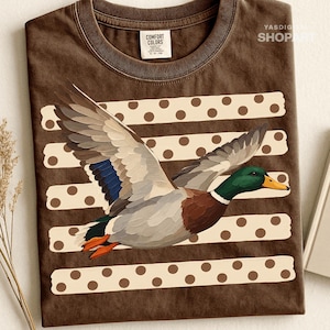 May include: A brown crew neck t-shirt featuring a graphic of a mallard duck in flight. The duck has a green head, yellow beak, and brown, grey, and blue wings. Behind the duck are three horizontal cream-coloured stripes with brown polka dots.