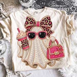 May include: A cream-colored t-shirt featuring a graphic of a ghost adorned with a large leopard print bow, pink-rimmed sunglasses, a leopard print iced coffee cup, and a bright pink handbag, all embellished with a sparkling, jewel-like texture.