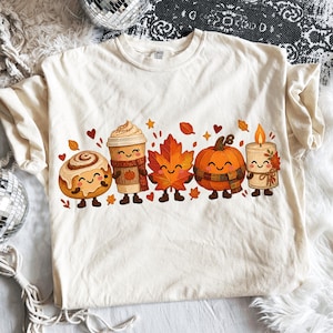 Puede incluir: Una camiseta de color crema con un estampado gráfico de cinco personajes otoñales sonrientes: un rollo de canela, una taza de café, una hoja de arce, una calabaza y una vela, rodeados de pequeñas hojas naranjas y rojas y corazones.