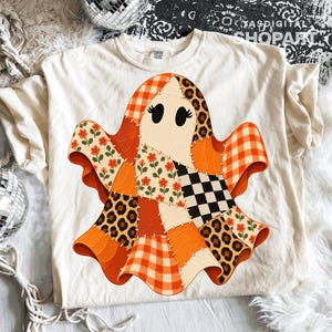 Może przedstawiać: Kremowy t-shirt z centralną grafiką ducha złożonego z różnych wzorów patchworkowych. Projekt ducha zawiera pomarańczową kratkę, wzór w panterkę, czarno-białą szachownicę oraz motywy kwiatowe, połączone widocznymi szwami na całej powierzchni.