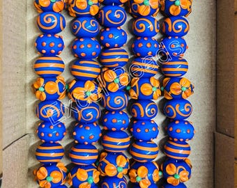 May include: Rows of vibrant blue beads with orange accents, including stripes, dots, and floral designs. The beads are strung together, ready for crafting. The beads are in a cardboard box.