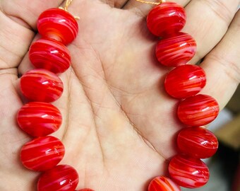 May include: A necklace of vibrant red beads with white swirling patterns. The beads are oval-shaped and strung together with a gold-colored cord. The necklace is held in a hand, showcasing the color and design of the jewelry.