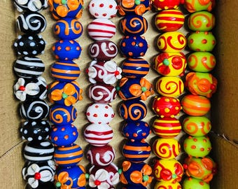 May include: A collection of colorful glass beads in a variety of designs. The beads feature stripes, dots, floral patterns, and swirls in shades of blue, orange, red, yellow, white, and green. The beads are strung together and displayed in a cardboard box.
