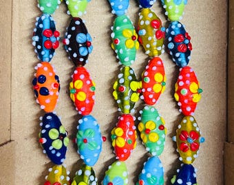 May include: A collection of colorful, handcrafted glass beads in various shades of blue, green, orange, and red. Each bead is oval-shaped and adorned with floral and polka dot designs. The beads are strung together with thin, natural-colored cords.