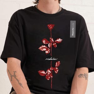 Puede incluir: Camiseta negra con un gráfico de rosa roja y la palabra "violator" en una fuente manuscrita. La camiseta tiene mangas cortas y cuello redondo. El texto "Depeche Mode" está impreso en una pequeña etiqueta en el lado derecho de la camiseta.