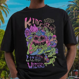 以下が含まれることがあります： 黒いTシャツ。カラフルなグラフィックデザインで、「King Lizard Wizard」と「Altered Beast」の文字と、ピンク、緑、青、紫の漫画の生き物が描かれています。ゆったりとしたオーバーサイズのフィット感です。