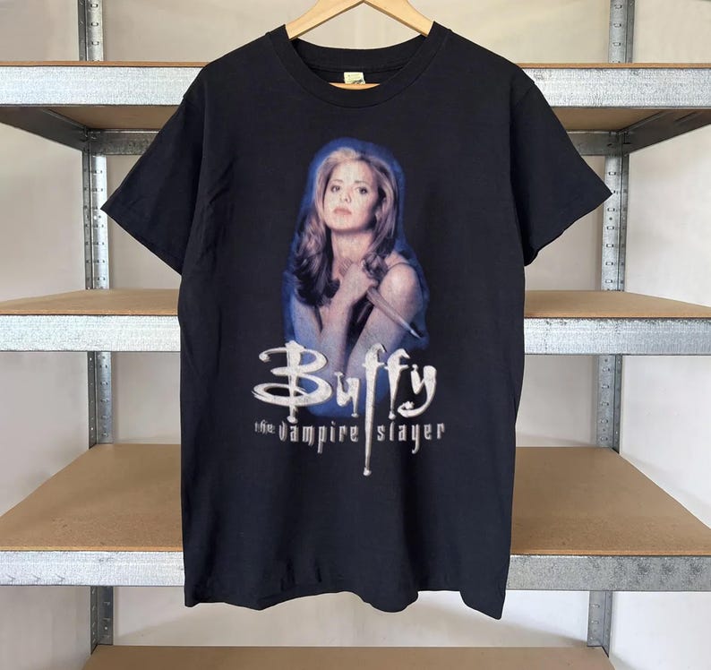 May include: Black t-shirt featuring a graphic of a woman with long blonde hair, holding a stake, with the text "Buffy the vampire slayer" in white. The shirt is hanging on a wooden hanger.