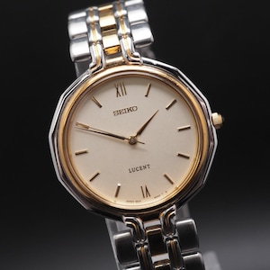 May include: A Seiko Lucent watch with a silver and gold-tone band and a cream-colored face. The watch face has gold-tone hands and Roman numeral hour markers. The watch is a classic design.