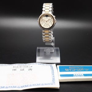 May include: A Seiko watch with a silver and gold-tone band and a cream-colored face. The watch is displayed on a clear stand. Included are a warranty document and a blue and white box with Japanese text.