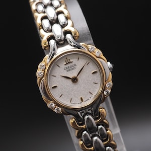 May include: A Credor Seiko wristwatch with a silver and gold-tone band and a white face. The watch has a round face with gold-tone accents and small diamonds. The watch band is made of silver and gold-tone links.