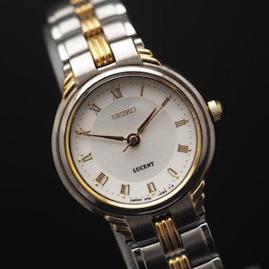 Könnte beinhalten: Eine Seiko Lucent Armbanduhr mit silbernem und goldfarbenem Metallband und Gehäuse. Das Zifferblatt ist weiß mit goldenen Zeigern und römischen Ziffern. Klassisches Design.