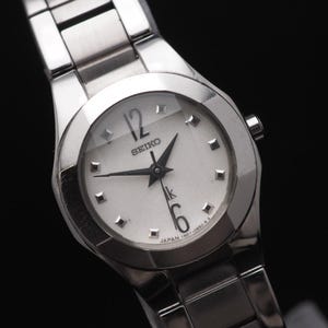 Può includere: Un orologio da polso Seiko argentato con quadrante bianco. L'orologio ha un cinturino argentato e un quadrante rotondo con lancette e indici neri. I numeri 12 e 6 sono visibili sul quadrante. L'orologio è su sfondo nero.