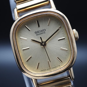 Può includere: Un orologio Seiko Quartz dorato con quadrante quadrato e cinturino dorato. Il quadrante è di colore oro chiaro con lancette e indicatori neri. L'orologio presenta la scritta "SEIKO QUARTZ".