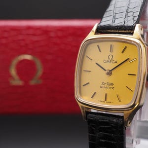 May include: An Omega De Ville quartz wristwatch with a gold-tone square case, a yellow dial, and a black textured leather strap. The dial features the Omega logo, the text Omega, De Ville, QUARTZ, and SWISS MADE. A red box with the Omega logo is visible.