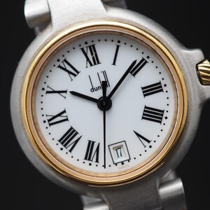 Puede incluir: Reloj de pulsera Dunhill con esfera blanca, bisel dorado y correa plateada. Cuenta con números romanos negros, manecillas negras y ventana de fecha a las 3. Diseño clásico y elegante.