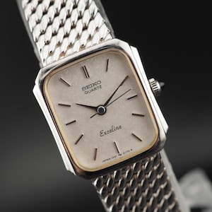 May include: A Seiko Quartz Exceline wristwatch with a rectangular silver-tone case, a gold-tone bezel, and a textured silver dial. The watch features black hands and hour markers, and a silver-tone metal mesh bracelet with a woven link pattern.