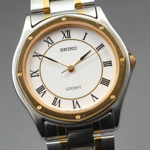 May include: A Seiko Lucent wristwatch featuring a white dial with black Roman numeral hour markers. The watch has a round gold-tone bezel with small studs and a two-tone stainless steel and gold-tone link bracelet with three thin hands.
