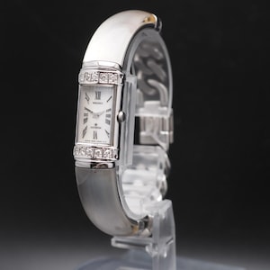 May include: A silver-toned wristwatch with a rectangular face and a diamond-studded bezel. The watch features a white dial with Roman numeral hour markers and a silver band. The watch is displayed on a clear stand against a dark background.