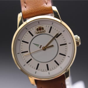 以下が含まれることがあります： 白文字盤、黒マーカー、金色の針を備えた金色のオリエント時計。 茶色の革バンドが付いています。 時計の文字盤にはオリエントのロゴとテキストがあります。 クラシックな時計です。