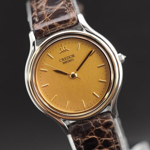 May include: A Credor Seiko wristwatch featuring a round gold-toned dial with thin gold hour markers and hands. The watch case is silver-toned metal with a gold-toned bezel. It is attached to a dark brown textured leather strap with a crocodile pattern.