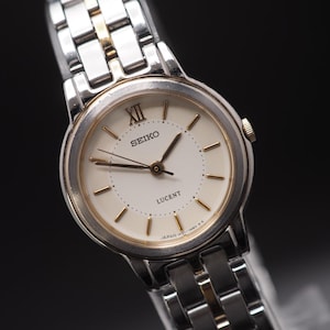 Op de afbeelding: Een close-up van een Seiko Lucent horloge met een zilveren en goudkleurige metalen band. De wijzerplaat is crèmekleurig met goudkleurige wijzers en markeringen. Het horloge heeft een klassiek ontwerp met Romeinse cijfer XII.