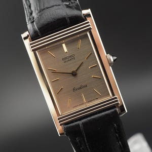 May include: A rectangular Seiko Quartz Exceline wristwatch with a gold-tone case, a brushed champagne dial featuring gold-tone hour markers and hands, and a black leather strap with a textured finish. The dial displays the text Seiko Quartz Exceline.