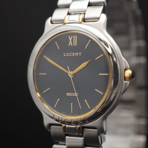 Könnte beinhalten: Eine silber- und goldfarbene Seiko Lucent Armbanduhr mit einem dunkelgrauen Zifferblatt. Die Uhr hat goldene Zeiger und Stundenmarkierungen sowie ein silbernes Armband. Das Wort "LUCENT" ist über der Mitte des Zifferblatts aufgedruckt.