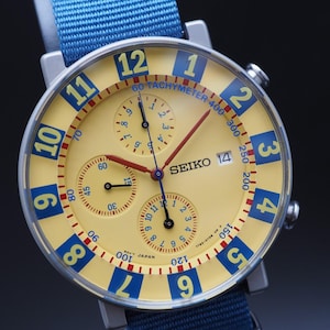 May include: A Seiko chronograph watch with a pale yellow dial, large blue Arabic numerals, and three sub-dials. The watch features a date window at the three o'clock position, red hands, and a blue nylon strap. The tachymeter scale is printed in blue.
