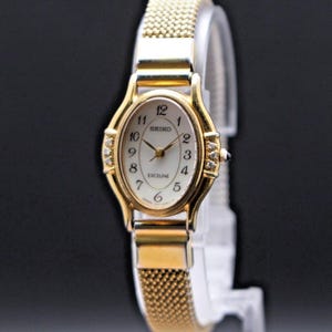 May include: A gold-toned Seiko Exciline wristwatch with a mesh band. The oval watch face has a white background with black numerals and hands. Small diamonds adorn the sides of the watch face.