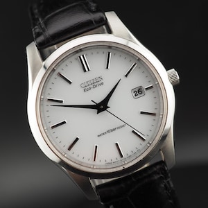 Op de afbeelding: Een Citizen Eco-Drive polshorloge met een ronde zilverkleurige roestvrijstalen kast, een witte wijzerplaat met zilverkleurige uurmarkeringen, zwarte wijzers, een datumvenster op de drie-uurpositie en een zwarte gestructureerde lederen band.