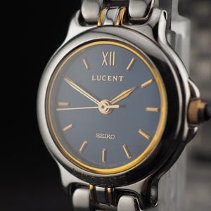May include: A two-tone wristwatch featuring a dark blue circular dial with gold-tone hour markers and hands. The watch face displays the text LUCENT and SEIKO, with a Roman numeral XII at the top. It has a silver and gold-tone metal link bracelet.
