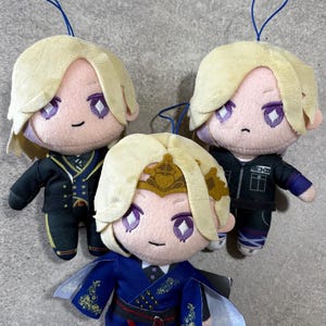 Puede incluir: Tres muñecos de peluche con cabello rubio y ojos morados, cada uno con un cordón azul para colgar. Un muñeco viste un traje azul estampado con una corona dorada, mientras que los otros dos llevan chaquetas negras con detalles y acentos azules.
