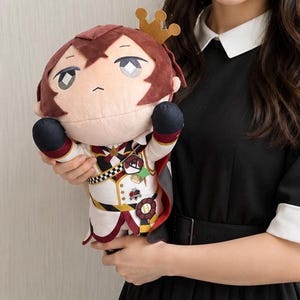 May include: A plush doll with a burgundy hair and a gold crown. The doll wears a white and black uniform with red accents and a cape. The doll is held by a person wearing a black dress.