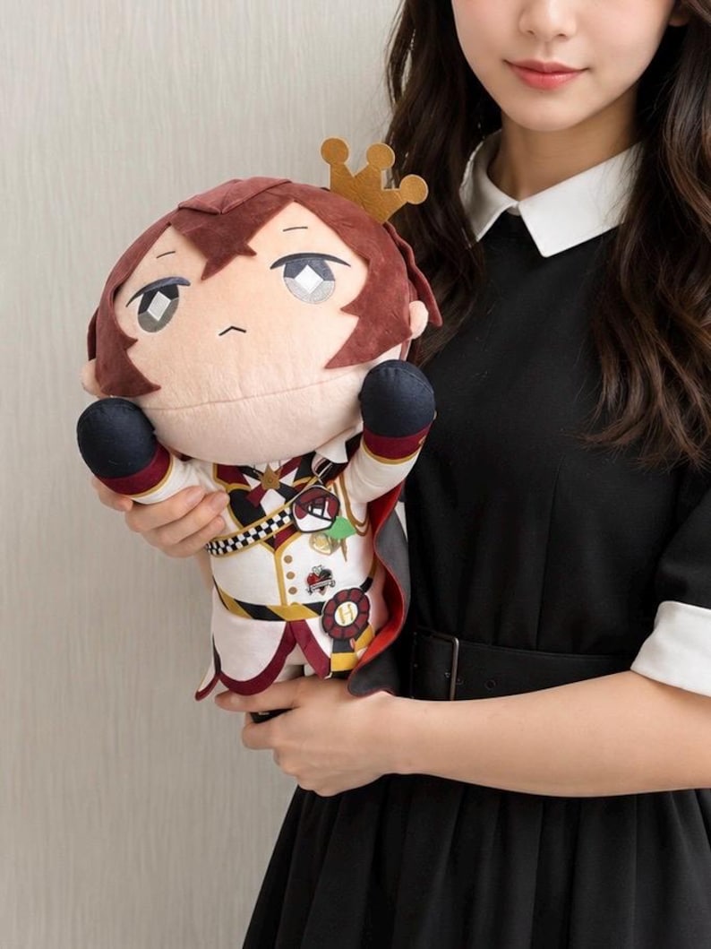 May include: A plush doll with a burgundy hair and a gold crown. The doll wears a white and black uniform with red accents and a cape. The doll is held by a person wearing a black dress.