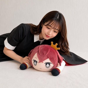 May include: A plush doll with a cartoon face, red hair, and a gold crown. The doll has a white and red outfit with black accents. The doll is lying on a white surface.