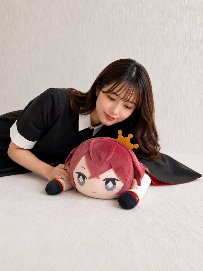 May include: A plush doll with a cartoon face, red hair, and a gold crown. The doll has a white and red outfit with black accents. The doll is lying on a white surface.