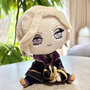 Puede incluir: Una muñeca de peluche con cabello rubio claro y ojos morados bordados. La muñeca viste un conjunto negro con cuello morado, ribetes dorados y patrones dorados en las mangas. Se muestra en posición sentada con pequeños pies visibles.