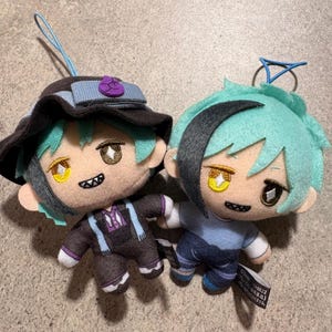 Può includere: Due bambole di peluche in stile anime con capelli verde acqua, una con cappello e completo neri, l'altra con camicia blu e jeans. Entrambe hanno occhi gialli e marroni e piccoli sorrisi. Ogni bambola ha un anello blu per appenderla.