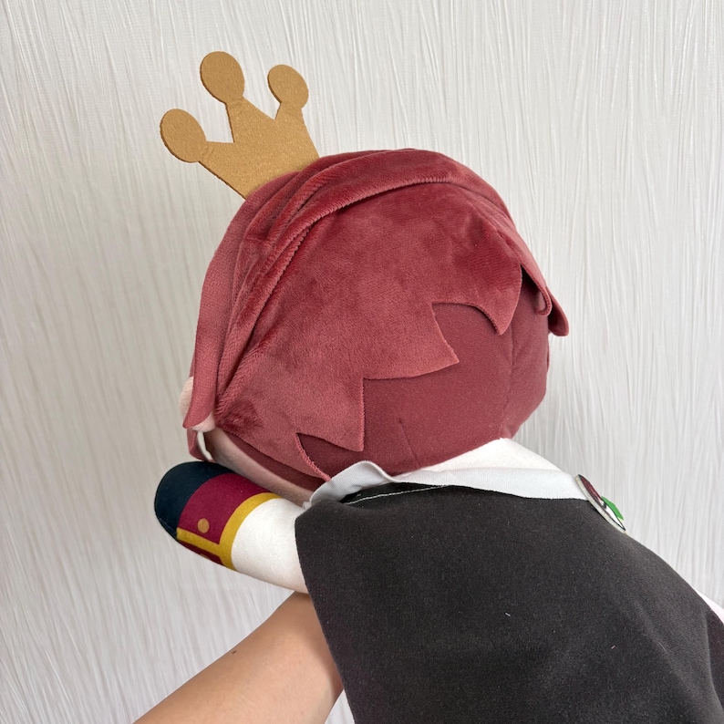 May include: A plush doll with burgundy hair and a gold crown. The doll is wearing a black cape over a white shirt. The doll's arm is visible, with a sleeve that has a red, gold, and black design.