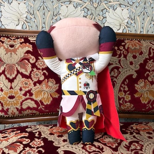 May include: A plush doll with a light pink head and a white uniform with red and black accents. The doll has its arms raised, and a red cape is draped over its back. The doll is decorated with gold and black details.