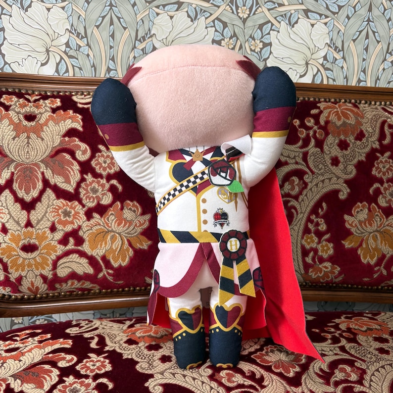 May include: A plush doll with a light pink head and a white uniform with red and black accents. The doll has its arms raised, and a red cape is draped over its back. The doll is decorated with gold and black details.