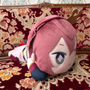 May include: A plush doll with pink hair and a detailed face, lying on a patterned red couch. The doll has a small, brown antler-like accessory and is wearing a detailed outfit with red, white, and gold accents. The doll is a collectible item.