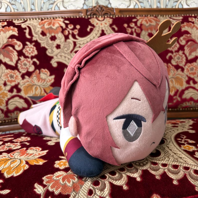 May include: A plush doll with pink hair and a detailed face, lying on a patterned red couch. The doll has a small, brown antler-like accessory and is wearing a detailed outfit with red, white, and gold accents. The doll is a collectible item.